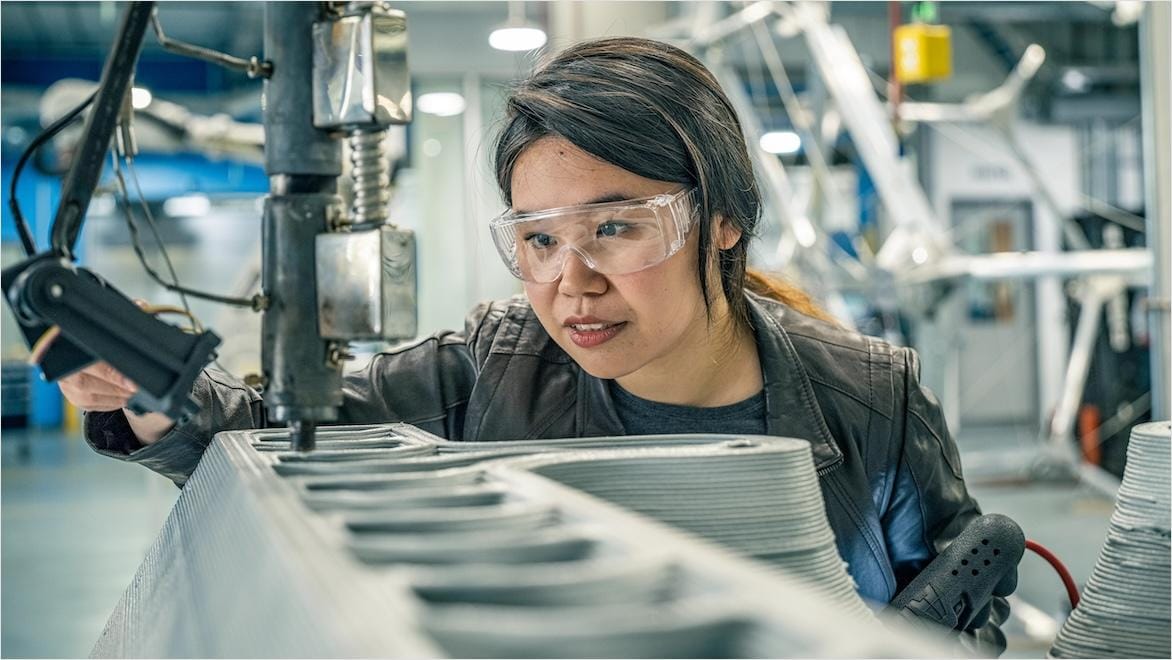 A woman wearing safety goggles adjusts the extrusion arm of a large 3D printing machine.