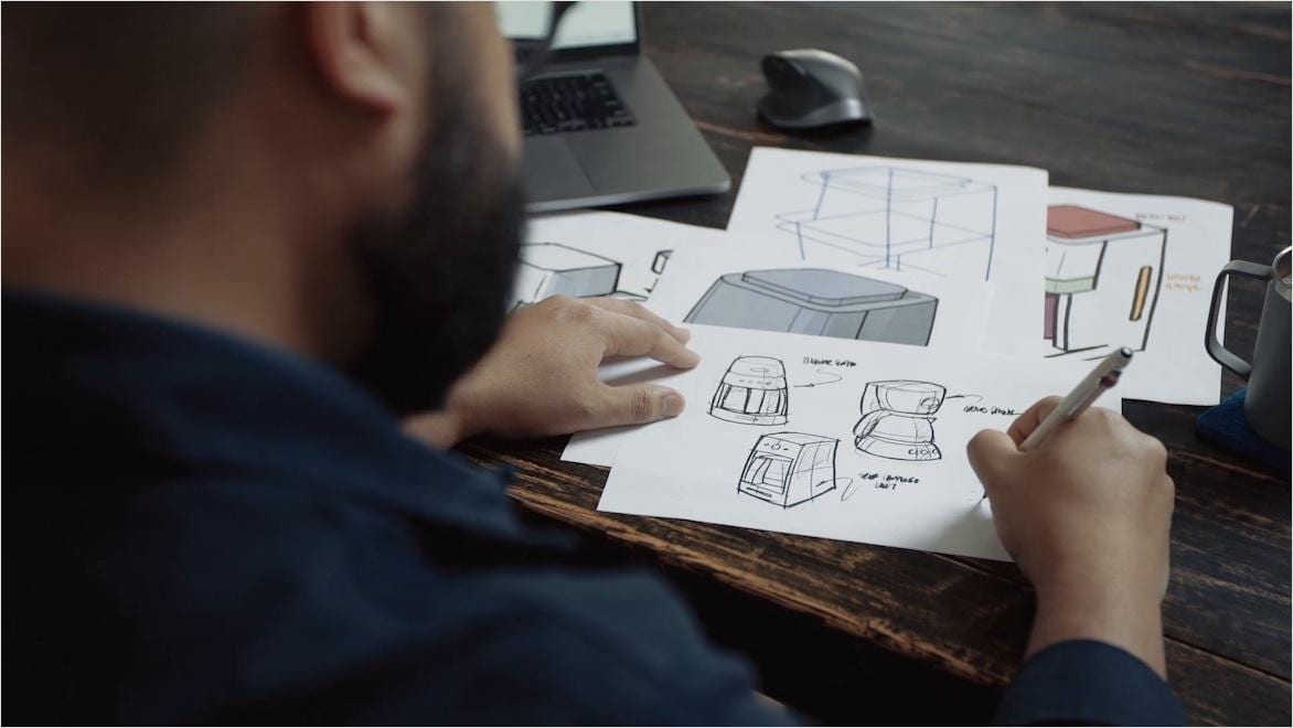 A view over the shoulder of a man sketching coffee maker designs on paper.