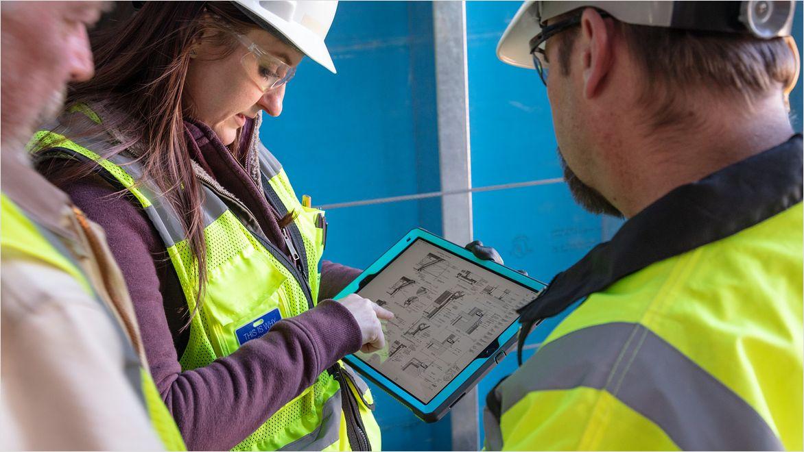 A woman and two men in safety gear on a construction site look at tablet the woman is holding that displays construction information.