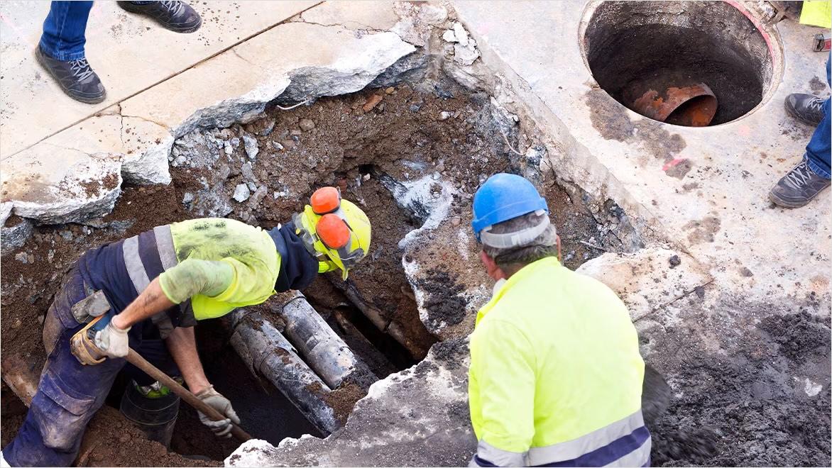 Workers in hard hats dig a hole to expose water pipes under a sidewalk.