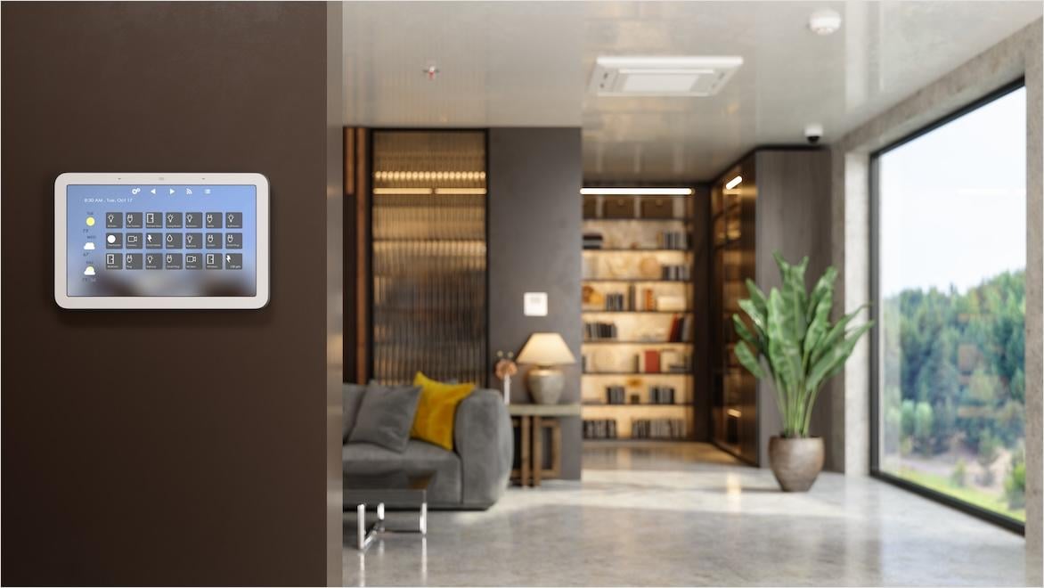 A screen with smart home controls is mounted on a wall next to the blurred background of a modern living room.
