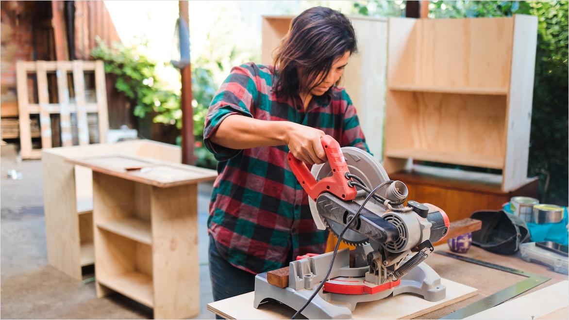 A woman uses a miter saw in her home wood shop; cabinet frames are visible behind her.