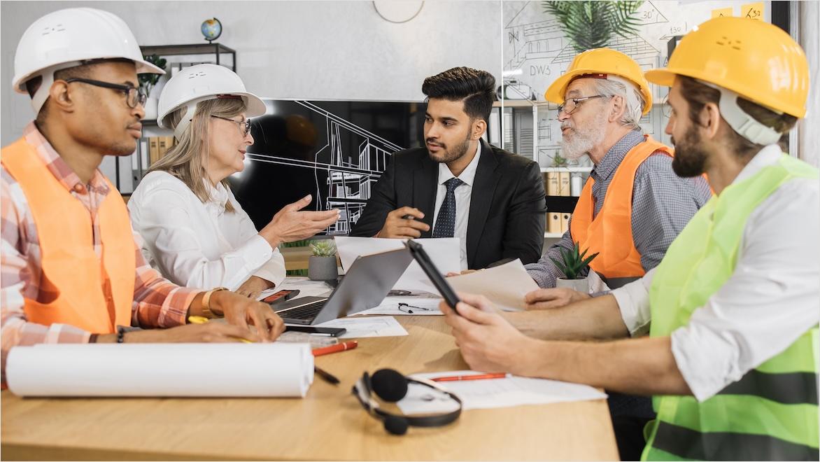 Five multiracial architects sit at a table in a modern office, reviewing a project.