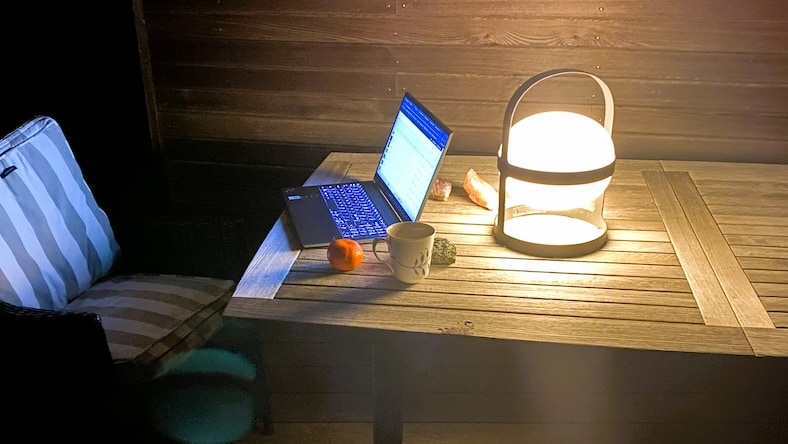 2170655838 A home office set up on an outdoor table after dark includes a laptop, mug, apple, deck chair, and round, glowing portable light.