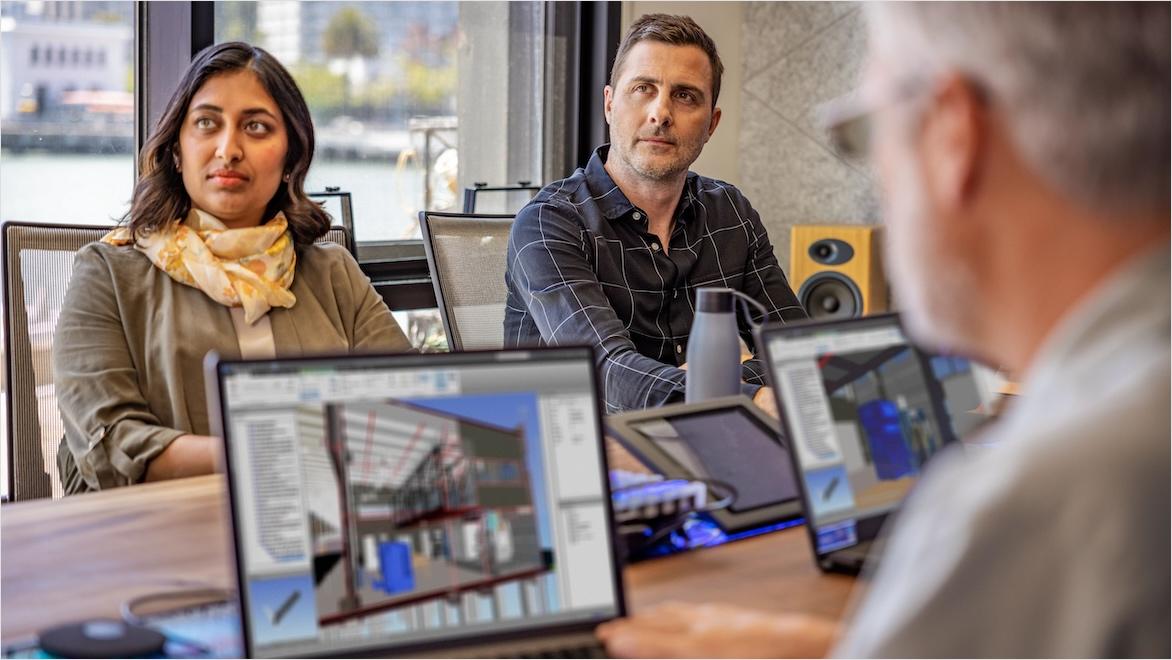 Several people sit around a conference table in a meeting; CAD software showing building designs is visible on several laptop monitors.