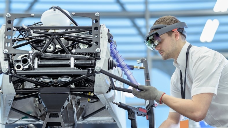 926846200 A man wearing a headset works on car parts in a factory; an overlay indicates the hologram he is viewing to help him position parts.
