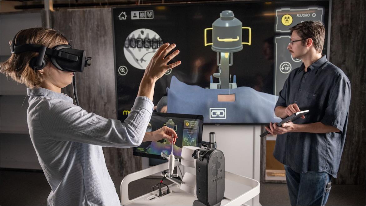A woman wearing a VR headset interacts with a VR and haptic feedback surgical training platform while her coworker watches.