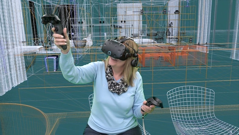A woman wearing a VR headset and using handheld controls is shown within a virtual architectural 3d wireframe environment.