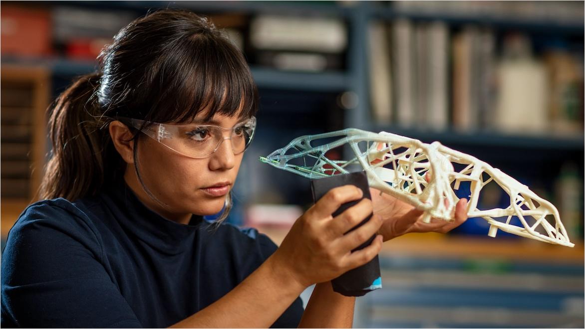 A woman wearing safety goggles holds a 3D-printed part created using generative design.