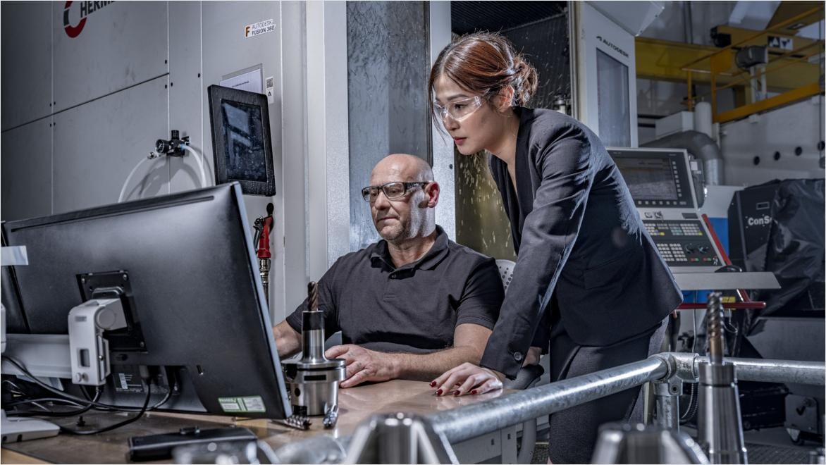 Two professionals collaborating in a manufacturing environment, with computer screens and CNC machinery in the background.