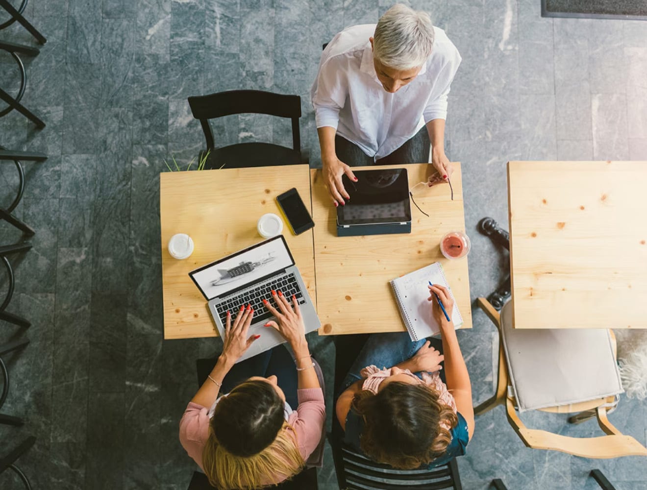 Três pessoas sentam-se em uma mesa trabalhando em laptops e fazendo anotações em notebooks.