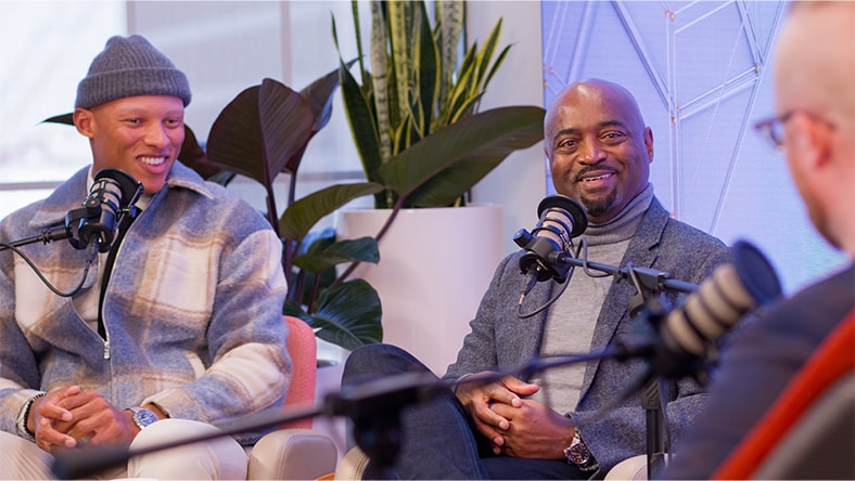 Three men sit in a podcast studio recording an episode, speaking into microphones. Two guests face the camera and smile while a third person is seen from behind in the foreground. Indoor plants and soft lighting create a modern, relaxed setting.