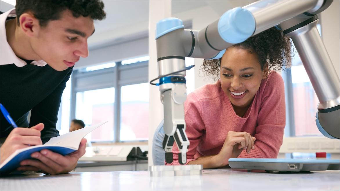 Two college students programming a robotic arm in a classroom.