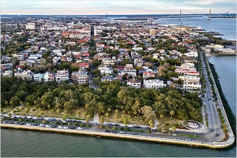 Aerial view of the city of Charleston's seawall, showing a coastal urban area with historic buildings, green spaces, and waterfront infrastructure.