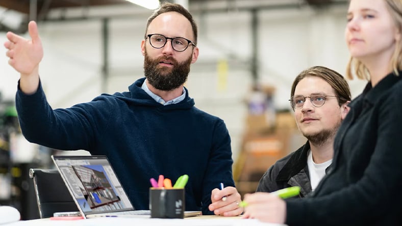 Illustration of two architects reviewing blueprints, one standing and one seated at a drafting table, with design elements and technical drawings visible around them.