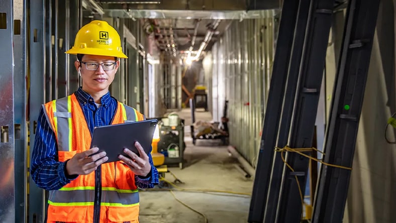 Illustration of two general contractors on a construction site, one reviewing plans on a tablet while the other observes, with cranes and building structures in the background.
