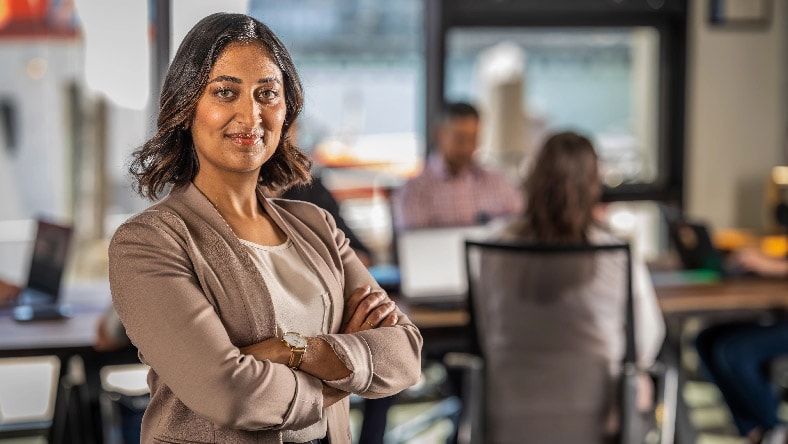 A smiling woman stands facing forward with her arms crossed, while coworkers sit at a conference table in the background.