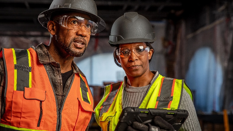 A man and a woman wearing safety vests, hard hats, and safety goggles confer over a tablet.