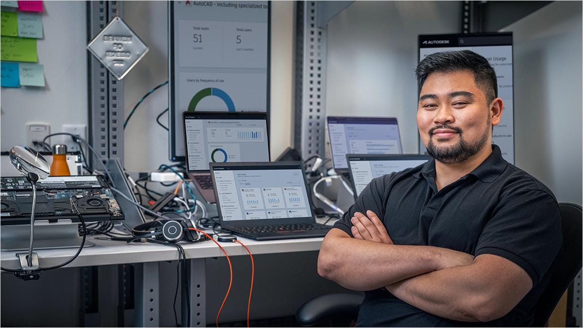 A smiling man sits with his arms crossed in front of several computer monitors.