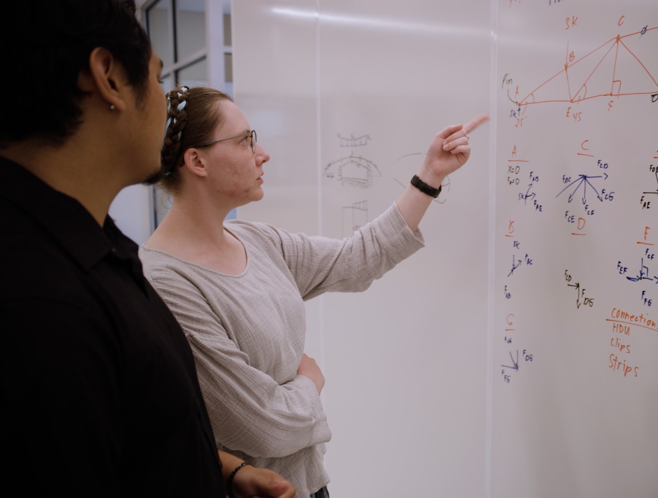 Two students stand at a large whiteboard covered with structural sketches, diagrams, and notes; one student points to a truss drawing while explaining a concept, and the other listens attentively.