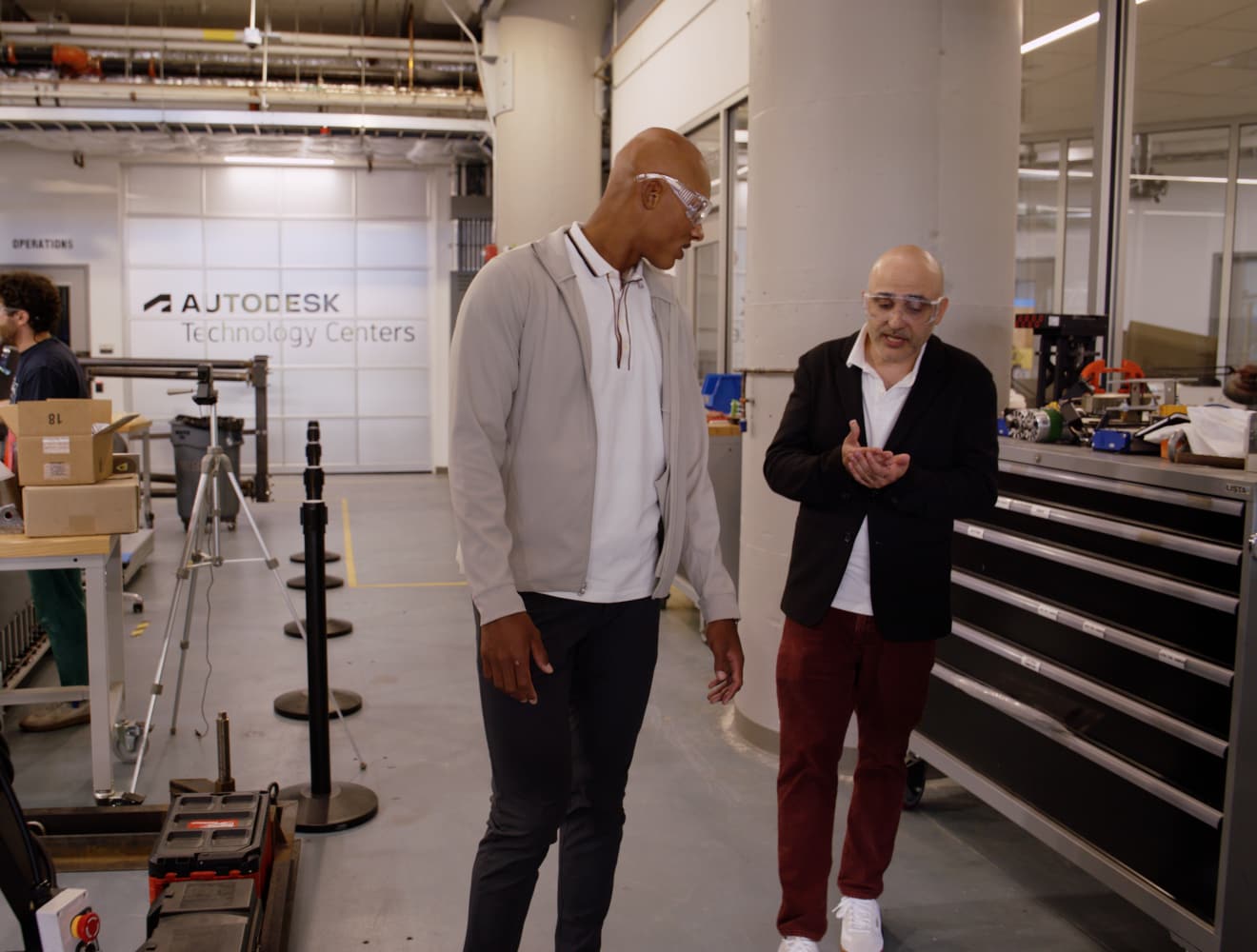 Josh Dobbs tours the Autodesk Technology Center with a staff member, both wearing safety glasses, while engineers work with equipment in the background.
