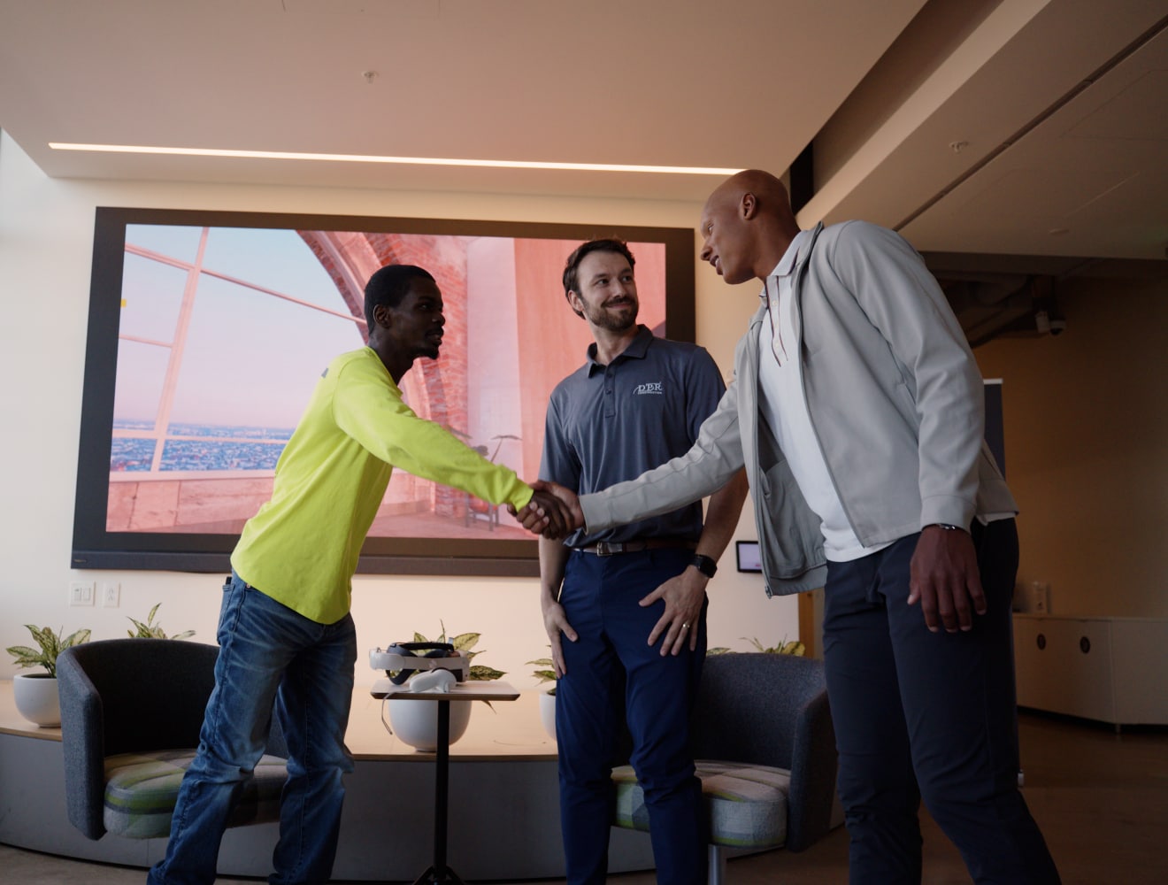 Josh Dobbs shakes hands with a young trades apprentice while a DPR Construction team member looks on, standing together in a bright lobby area with a large screen behind them.