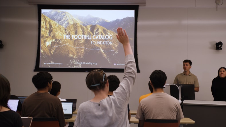 A student raises their hand during a classroom presentation, while two speakers stand at the front near a screen displaying “The Foothill Catalog Foundation” over an image of mountain terrain.