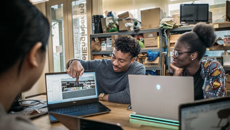 Three students sit around a table with laptops, while one points to a digital building design on his screen and discusses it with the group in a workshop-style classroom.