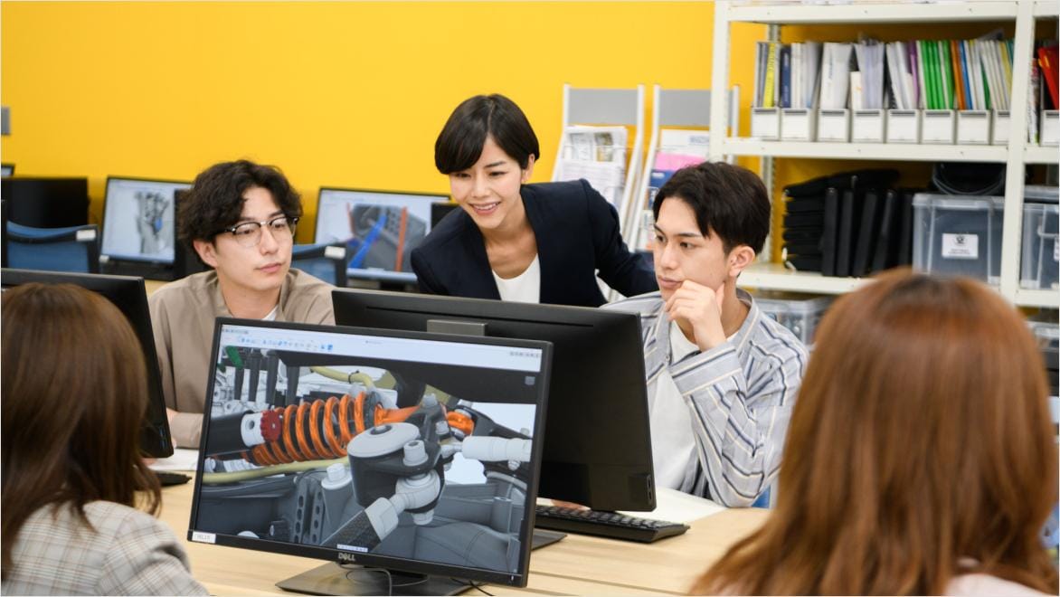 Several student's work on a project on desktop, their teacher leans forwards and shows two students something on their screen. The student's are working in Autodesk Fusion. 