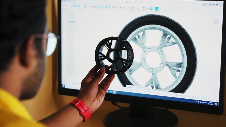 A man with in glasses holds a wheel up to a desktop screen displaying the wheel's design in Autodesk Fusion, we see only the back of the man's head