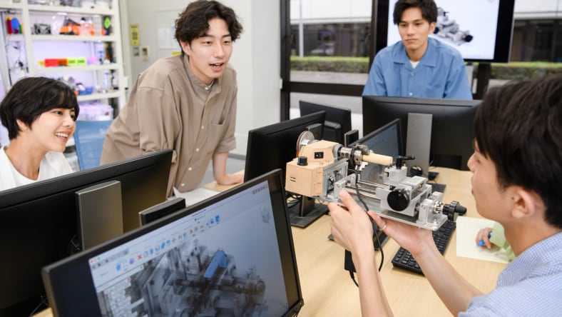 Four students lean over to look at a robot build, they are working in Autodesk Fusion on their desktops