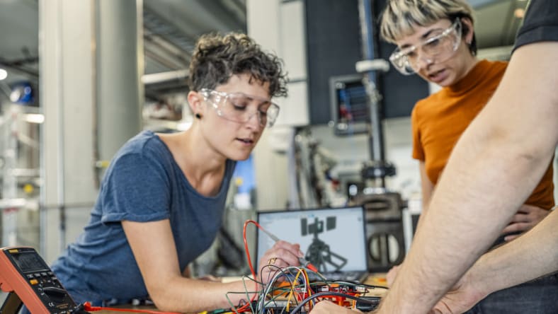 Two women work on an electronics project using Autodesk Fusion, both are wearing safety goggles