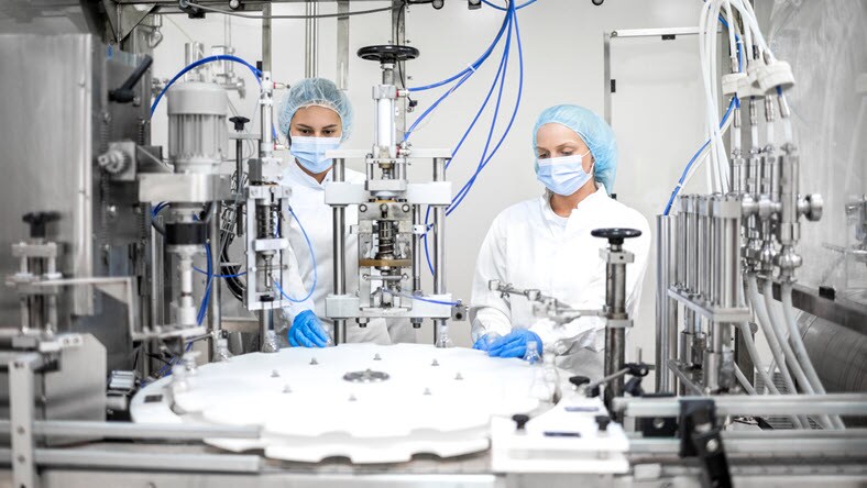 Due giovani operatrici con dispositivi di protezione azionano una macchina farmaceutica di alta tecnologia che esegue più funzioni, tra cui soffiare, riempire, tappare ed etichettare bottiglie.