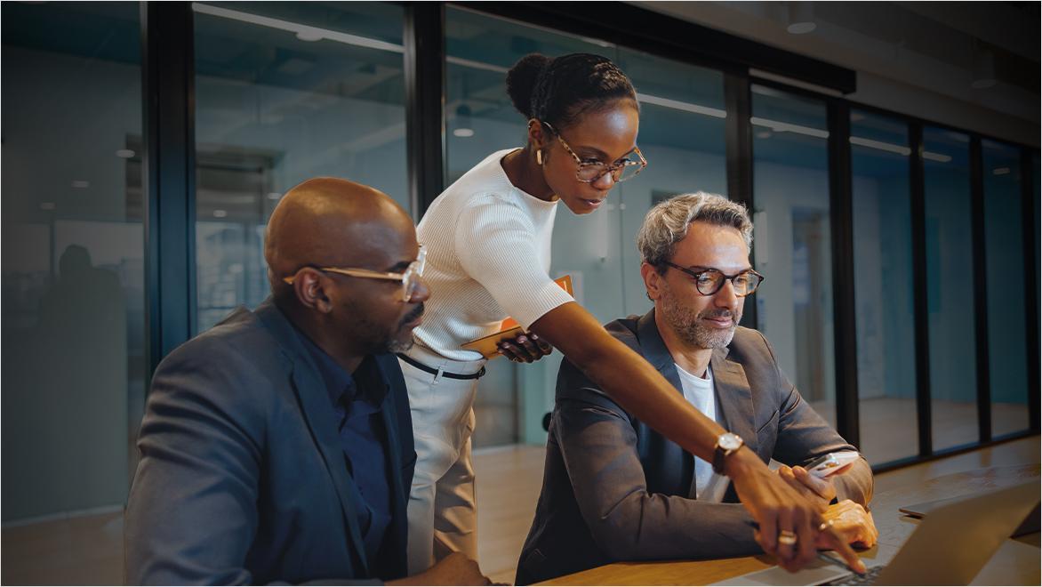 Three business professionals collaborating around a laptop in a modern office with glass walls. A woman in a white top stands between two seated men in business attire, pointing at the laptop screen while they review content together.