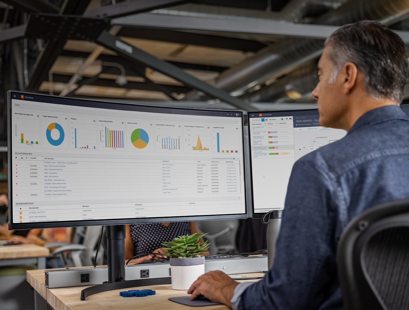A man seated at a desk, working on two computer monitors displaying a dashboard.