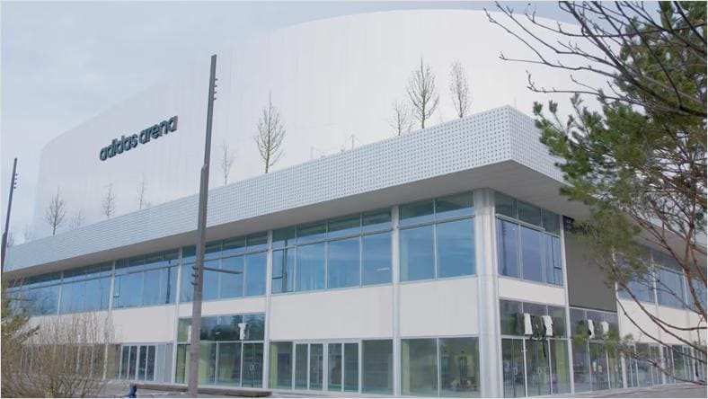 Vue extérieure de l'adidas arena, bâtiment moderne avec de grandes fenêtres, arbres sur le toit, et le nom 'adidas arena' en lettres noires.