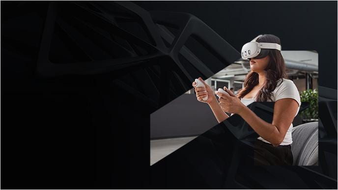 A woman wearing a virtual reality headset sits indoors and uses handheld controllers.