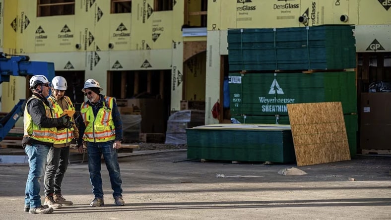 Three construction workers in hard hats review plans at an active building site with stacked materials behind them.