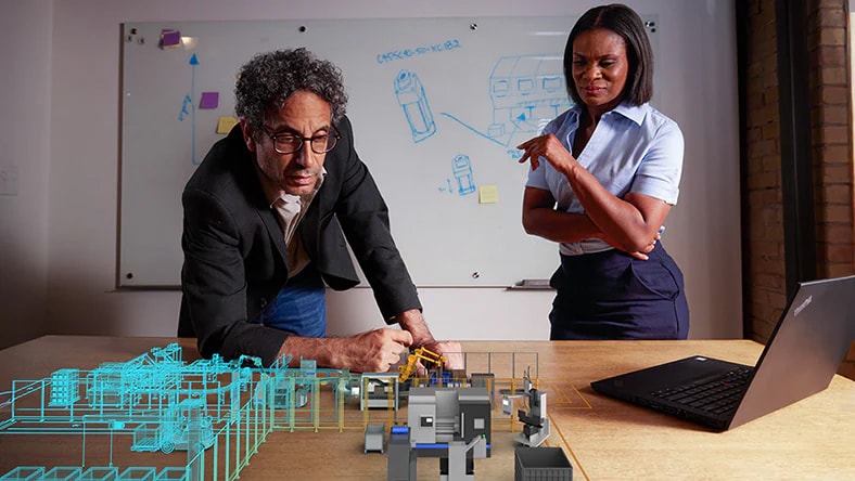 A man and a woman stand in an office. Before them is a desk with a laptop on it, next to the laptop there's a digital representation of a factory next to a factory model. They look at the model with a focused facial expression.