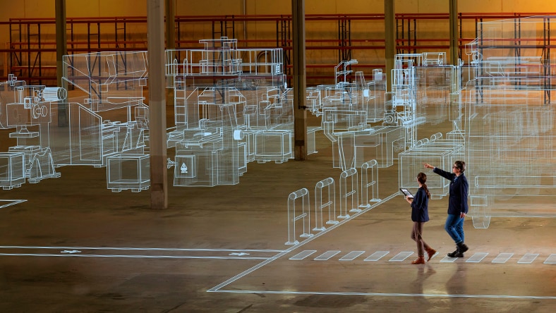 Two people walking through an industrial warehouse space overlaid with a glowing wireframe digital twin visualization of factory equipment and structures, illustrating digital factory technology.