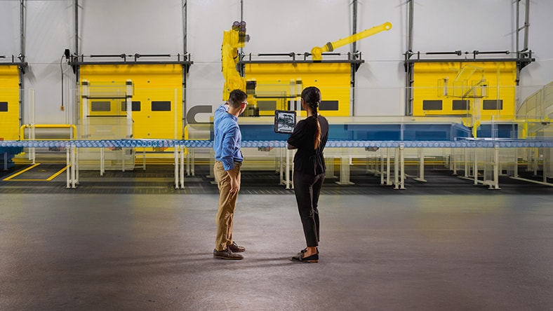 Two people standing beside a large yellow industrial robotic arm in a workshop, looking at a computer screen displaying a 3D model, suggesting collaboration in engineering or automated manufacturing.