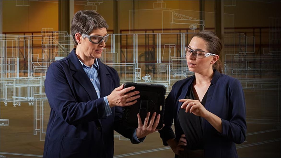 Two people in business attire discussing while holding a tablet with a transparent architectural blueprint overlay in the background.