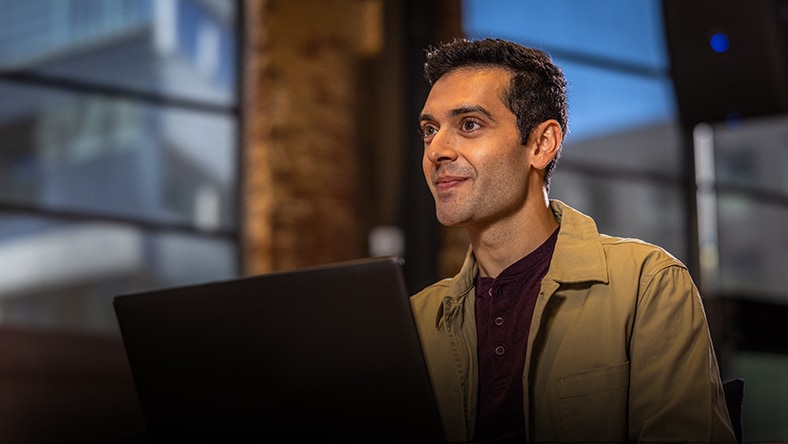 Person with short dark hair wearing a tan jacket and dark shirt sitting behind a laptop in an indoor setting with blurred background.