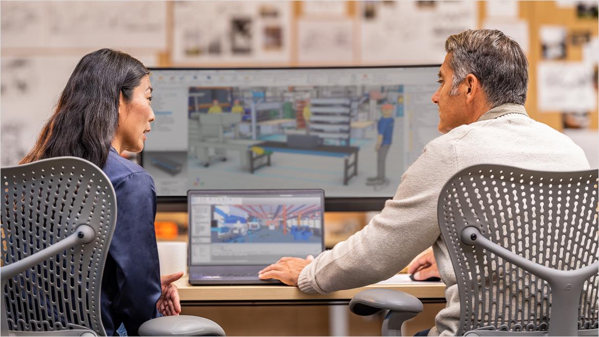 Two people sit at desks, looking at a laptop and a large monitor displaying 3D design software with industrial interior renderings.