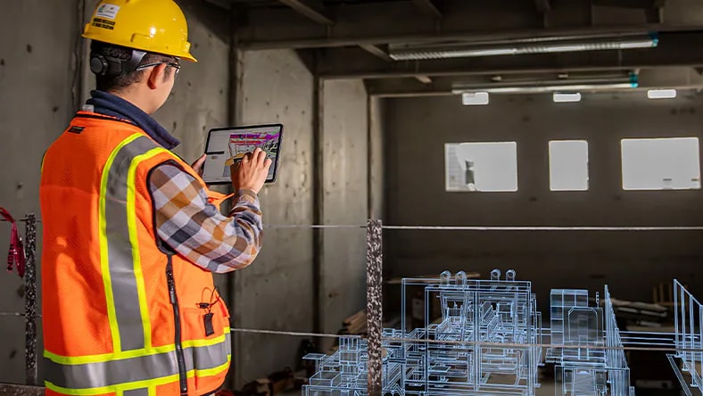 A man wearing a hard hat and a safety vest uses an iPad to configure a digital representation of a factory in front of him.