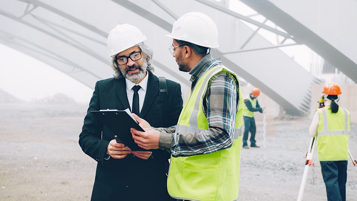 Two men wearing white safety helmets and high-visibility vests talking at a construction site with other workers in the background