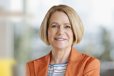 Smiling woman with short blonde hair wearing an orange blazer and striped shirt against a blurred background