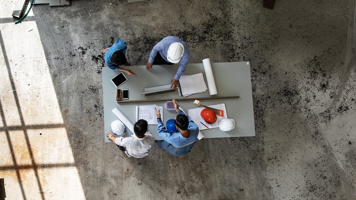 Four people wearing hard hats gathered around a table with blueprints and a tablet on a concrete floor