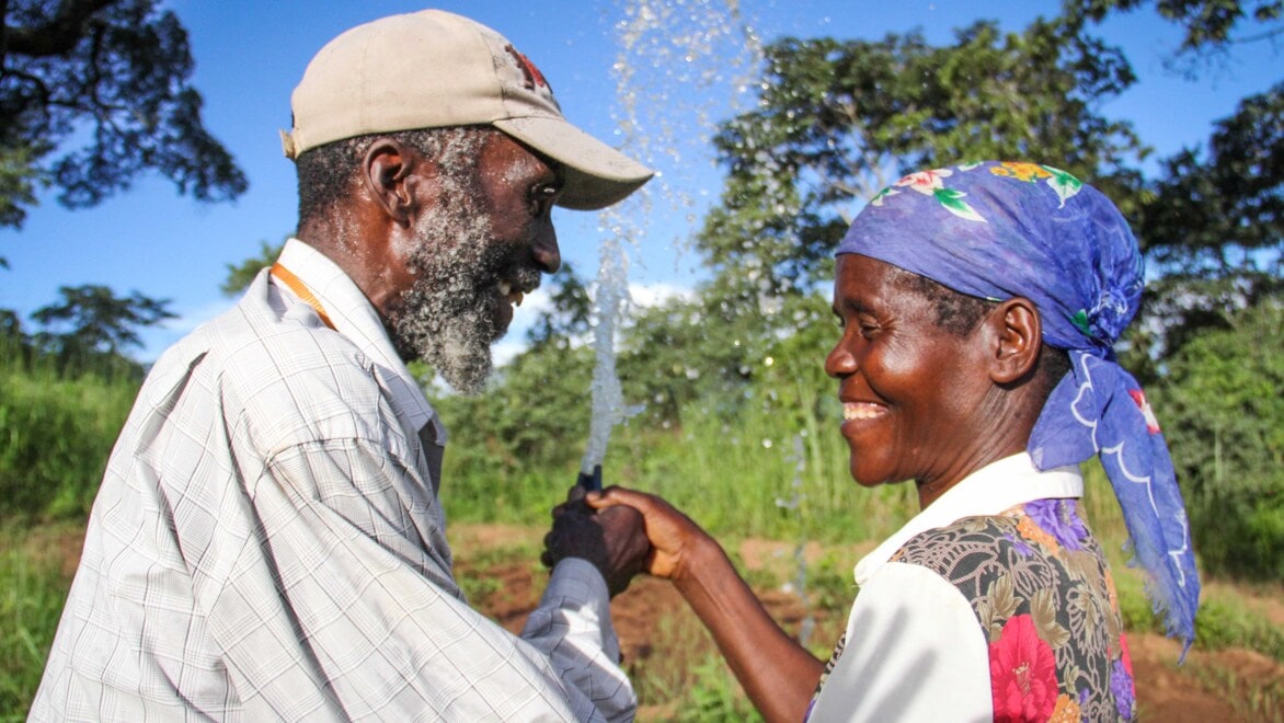 A man wearing a beige baseball hat and woman wearing a purple scarf, holding a hose spraying water into a garden, standing outside, smiling and looking at each other.