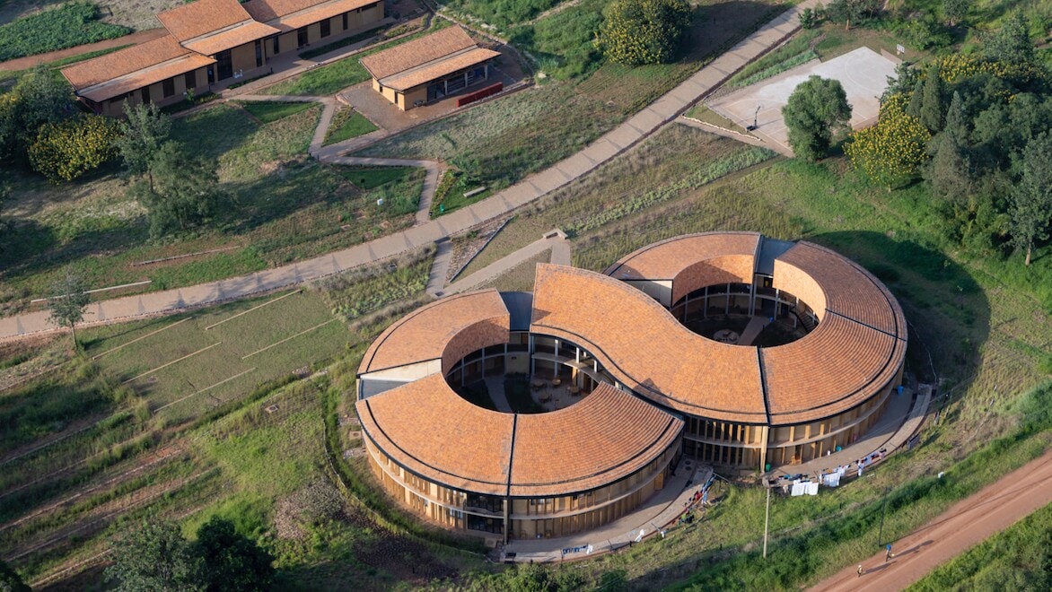 Aerial view of MASS Design Group's Rwanda Institute for Conservation Agriculture (RICA). Building shaped like an infinity symbol, surrounded by rural land and other small buildings.